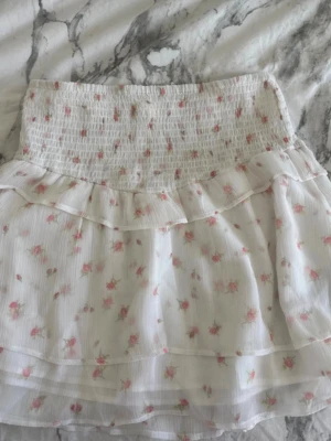 Vit blommig volangkjol Gina Tricot - Supersöt vit kjol från Gina Tricot med små rosa blommor och volangdetaljer. Kjolen har smock i midjan för skön passform och är gjord i ett lätt, luftigt material. Perfekt för sommarens alla dagar!