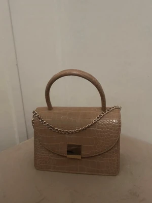 Beige handväska med krokoprägling - Snygg beige handväska med krokodilpräglat mönster och guldfärgat spänne. Väskan har ett stadigt handtag och en silverfärgad kedja som axelrem. Perfekt accessoar för att lyfta din outfit.
