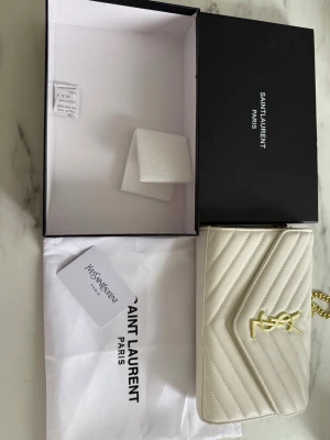 Vit kuvertväska från Saint Laurent - Snygg vit kuvertväska från Saint Laurent med klassiskt YSL-emblem i guld framtill. Väskan är i quiltat läder med kedjerem i guld och magnetlås. Kommer med originalbox, dustbag och äkthetskort. Perfekt accessoar för att lyfta din outfit.