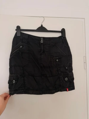 Svart utility minikjol med fickor - Cool svart kortkjol med flera fickor, dragkedja och knappar. Utility-stil med markerade sömmar och detaljer. Perfekt för en edgy streetlook. Liten röd etikett på sidan. Passar dig som gillar praktiska detaljer och stil.