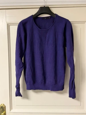 Lila stickad tröja från Åhléns - Snygg lila stickad tröja från Åhléns i 100% ekologisk bomull. Tröjan har rund halsringning, långa ärmar och ribbade muddar vid ärmslut och nederkant. Perfekt för dig som gillar stilrena och bekväma plagg.