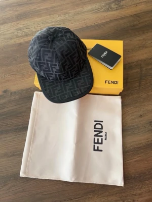 Fendi keps med FF-mönster - Snygg keps från Fendi med klassiskt FF-logomönster i mörkgrått och svart. Kepsen har böjd skärm och justerbar baksida. Materialet är textil och kepsen levereras med originalbox och dustbag. Perfekt för dig som vill ha en exklusiv accessoar.