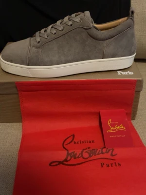 Gråa sneakers från Christian Louboutin - Snygga gråa sneakers från Christian Louboutin i mocka med vita sulor och klassisk låg profil. Skorna har diskreta detaljer och stilrena snörningar, perfekt för dig som vill ha en exklusiv look. Kommer med originalkartong och röd dustbag.