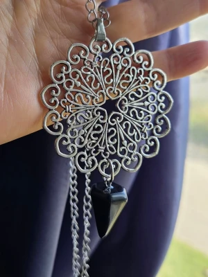 Halsband med silverfärgat mönster goth alternative gothic mandala indisk  - Unikt halsband med en stor, rund och detaljerad silverfärgad medaljong i filigran-design. Hängsmycket har en svart, konformad sten som ger en cool kontrast. Kedjan är i metall och har ett klassiskt lås. Perfekt för dig som gillar statement-smycken och snygga detaljer. Material: glassten och hänge av zinklegering. Hänget är 8x5 cm. 