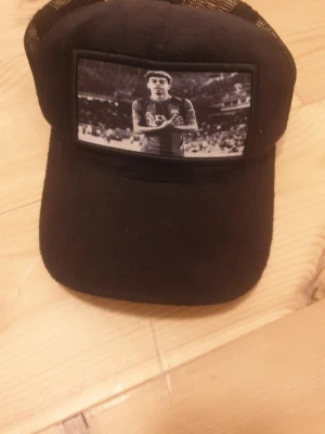 Svart keps från Evosportswear - Svart keps från Evosportswear med mesh baktill och en stor svartvit patch framtill med motiv av en fotbollsspelare. Justerbar passform och sportig design, perfekt för dig som gillar streetstyle och sportmode.
