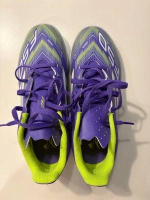 Adidas F50 lila fotbollsskor - Säljer ett par Adidas F50 fotbollsskor i lila med neongula detaljer och vita ränder. Skorna har snörning, låg profil och gummisula som passar perfekt för inomhusspel. Snygg design med grafiskt mönster och bekväm vadderad insida. 
