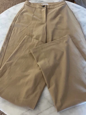 Beige kostymbyxa från Bershka - Stilrena beige kostymbyxor med raka ben och pressveck framtill. Byxorna har en klassisk passform, dold dragkedja och knapp i midjan samt en bakficka med knapp. Perfekta för en clean och uppklädd look. Byxan har svarta färgskiftningar och små svarta prickar som bilden visar, dock ej så synliga.