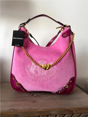 Juicy Couture rosa axelväska - Snygg rosa axelväska från Juicy Couture i sammet med mörkrosa läderdetaljer. Väskan har en guldfärgad kedja med hjärtdetalj och präglad logga framtill. Perfekt för dig som vill sticka ut med en trendig och färgglad accessoar.