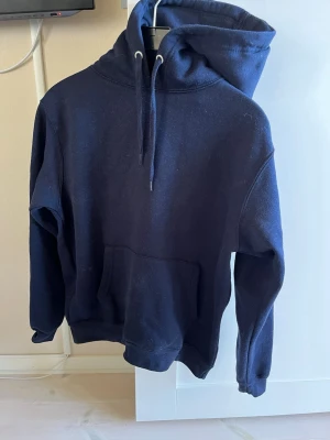 Mörkblå hoodie med ficka - En enkel och stilren mörkblå hoodie med stor huva och dragsnören. Hoodien har en klassisk känguruficka framtill och ribbade muddar vid ärmslut och nederkant. Perfekt för en avslappnad och cool look.mörkblå