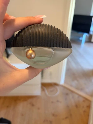 Kenzo Amour parfymflaska - Kenzo Amour parfym i en unik, halvmåneformad flaska med frostat glas och svart räfflat lock. Flaskan har en dekorativ guldfärgad pärla som detalj. Perfekt för dig som gillar snygg design och vill ha något annorlunda på parfymhyllan.