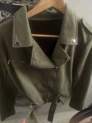 Olivgrön mocca jacka med bälte - Snygg olivgrön mocca jacka med klassisk bikerkrage, silverfärgade dragkedjor och knappar samt bälte i midjan. Jackan har axelklaffar och en cool, tidlös look som passar perfekt till jeans eller klänning.