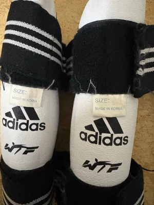 Adidas taekwondo skydd och handskar - Säljer ett set med vita och svarta taekwondo skydd och handskar från Adidas, märkta med WTF. Skydden har kardborreband och handskarna har förstärkta partier för extra skydd. Perfekt för kampsportsträning.