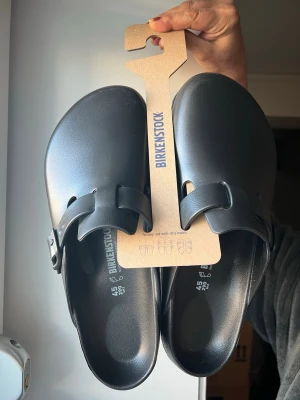Svarta Birkenstock Boston sandaler - Säljer ett par svarta Birkenstock Boston sandaler i EVA-material. De har en klassisk clog-design med justerbart spänne på sidan och en bekväm, formad sula. Perfekta för dig som gillar stilrena och praktiska skor.