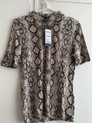 Beige topp med ormmönster New Yorker - Trendig topp från New Yorker i beige och svart med ormmönster. Modellen har korta ärmar och en liten ståkrage. Perfekt för dig som vill sticka ut med ett djurmönstrat plagg. Materialet är mjukt och stretchigt för en bekväm passform.