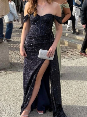 Glittrig marinblå balklänning - Snygg marinblå långklänning med glittrigt tyg och offshoulder-design. Klänningen har en markerad midja, slits framtill och en elegant v-ringning. Perfekt för bal eller festliga tillfällen.