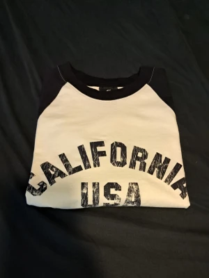 Svartvit t-shirt California USA - Cool svartvit t-shirt från H&M med trycket 'California USA' på bröstet. T-shirten har svarta ärmar och krage, medan kroppen är vit. Perfekt för en avslappnad och sportig stil.