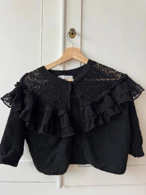 Svart kofta med volang från Zara - Svart kofta från Zara med snygga virkade detaljer och volanger över axlarna. Storlek S, fing skick!
