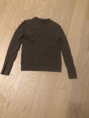 Grå kabelstickad tröja Morris - Kabelstickad grå tröja från Morris i 100% merinoull. Tröjan har rund halsringning, långa ärmar och en röd broderad logotyp på bröstet. Perfekt för dig som gillar klassisk och stilren design.