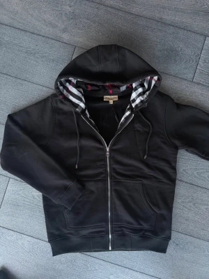 Svart hoodie med dragkedja från Burberry - Snygg svart hoodie från Burberry med dragkedja framtill och klassiskt rutigt foder i huvan och längs insidan. Tröjan har två stora fickor fram, justerbar huva med snören och ribbade muddar. Perfekt för en stilren och avslappnad look.