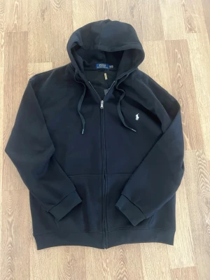 Svart hoodie från Polo Ralph Lauren - Svart hoodie med dragkedja från Polo Ralph Lauren. Klassisk design med huva och dragsko, samt den ikoniska vita broderade loggan på bröstet. Mjuk bomullskänsla och ribbade muddar vid ärmslut och nederkant. Perfekt för en avslappnad och stilren look.