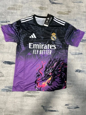 Real Madrid x Adidas draktröja lila - Snygg Real Madrid-tröja från Adidas i svart och lila med coolt drakmotiv och klubbmärke på bröstet. Tröjan har korta ärmar, rund hals och vita ränder på axlarna. Materialet är lätt och andas, perfekt för träning eller match.