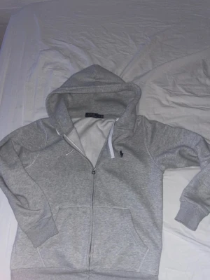Grå hoodie från Polo Ralph Lauren - Snygg grå zip-hoodie från Polo Ralph Lauren med klassisk huva och dragsko. Mjuk bomullsblandning, två fickor fram och ikoniska loggan broderad på bröstet. Perfekt för en chill och stilren look.