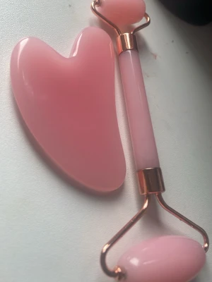 Rosa ansiktsroller och gua sha-set - Ett set med en ansiktsroller och en gua sha-sten i ljusrosa sten, perfekt för ansiktsmassage och hudvård. Rollern har metallfästen i roséguld och två rullar i olika storlekar. Gua sha-stenen har en hjärtformad design.