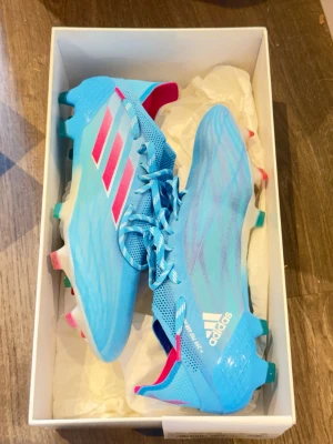 Adidas X Speedflow blå fotbollsskor - Snygga Adidas X Speedflow fotbollsskor i ljusblått med rosa och vita detaljer. Skorna har snörning, låg profil och är tillverkade i lätt syntetmaterial för maximal fart på planen. Perfekta för dig som vill sticka ut och spela snabbt.