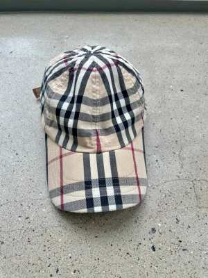Burberry keps  - Säljer en klassisk keps från Burberry,   i beige med det ikoniska rutmönstret i svart, vitt och rött. Kepsen har böjd skärm och justerbar rem bak. Tillverkad i bomull för en bekväm och stilren look.