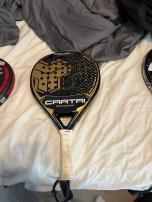 Cartri Cannon 3.0 padelracket - Snyggt padelracket från Cartri, modell Cannon 3.0, i svart och guld med vit lindning på handtaget. Racketen har rund form och är tillverkad i kompositmaterial med kolfiberstruktur. Perfekt för dig som vill ta ditt padelspel till nästa nivå.