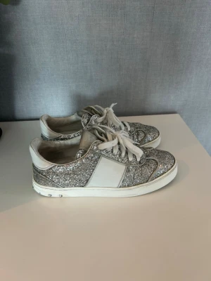 Valentino flycrews  - Säljer ett par riktigt snygga sneakers med massor av silverglitter och vita paneler. Skorna har vita snören och en vit sula med små metallnitar på hälen. Perfekta för dig som vill sticka ut och addera lite bling till din outfit. 4 nitar saknas samt limmat på något ställe