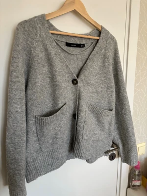 Grå kofta från Vero Moda - Mysig grå kofta från Vero Moda med v-ringning och tre knappar framtill. Koftan har två stora fickor.