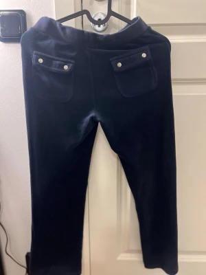 Mörkblå juicy couture mjukisbyxor med fickor - Mörkblå mjukisbyxor med snörning i midjan och stora fickor både fram och bak. Byxorna har silverfärgade detaljer på fickorna och snörningen. Materialet är mjukt och skönt, perfekt för chill dagar. Passformen är avslappnad och benen är raka.