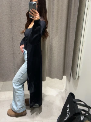 Lång svart cardigan med knappar - Snygg lång svart cardigan med knäppning framtill. Modellen är rak och har långa ärmar, perfekt att slänga över en topp eller t-shirt. Enkel och stilren design som passar till många outfits.