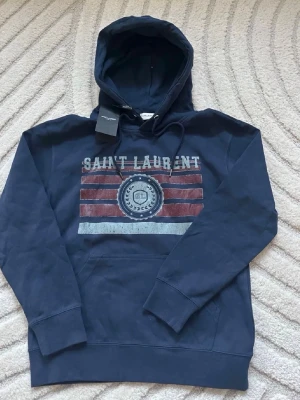 Mörkblå Saint Laurent hoodie - Snygg mörkblå hoodie från Saint Laurent med tryckt logga och ränder i rött och vitt på bröstet. Klassisk huva med dragsko och känguruficka framtill. Perfekt för en avslappnad och trendig stil. Storlek M väldigt bra skick!