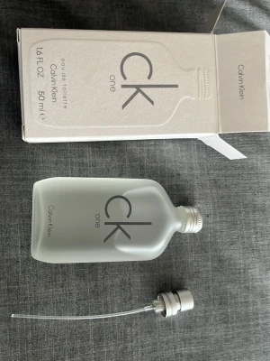 CK One Eau de Toilette 50ml - CK One från Calvin Klein är en ikonisk unisexparfym i en stilren, frostad glasflaska med minimalistisk design. Doften är fräsch och citrusaktig med inslag av gröna och träiga noter. Flaskan rymmer 50 ml och har en enkel, modern look med grå text.             Den är oöppnad och rätt så ny.
