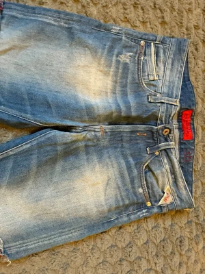 Blå slitna jeans från Replay - Säljer ett par blå jeans från Replay med slitningar och tvättade detaljer för en cool, avslappnad look. Klassisk femficksmodell med gylf och knapp, samt Replay-logga på fickan och röd etikett i midjan. Perfekta för dig som gillar streetstyle.