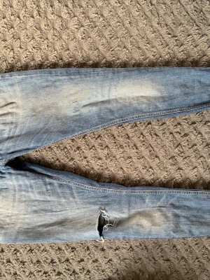 Ljusblå slitna jeans från Nudie - Säljer ett par ljusblå jeans från Nudie med skinny fit och slitningar, inklusive en större reva på ena benet. Jeansen har klassisk femficksdesign, gylf med dragkedja och är tillverkade i bomullsdenim. Perfekta för en avslappnad streetstil.