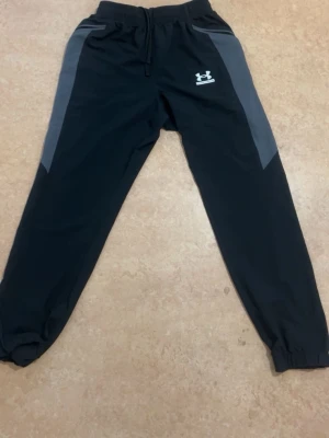Svarta mjukisbyxor från Under Armour - Snygga svarta mjukisbyxor från Under Armour med grå sidopaneler och resår i midjan samt benslut. Byxorna har dragsko i midjan och logga på vänster lår. Perfekta för chill eller träning.