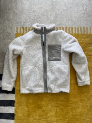 Vit teddyjacka från JJ Studios - Mysig vit teddyjacka från JJ Studios med grå detaljer. Jackan har hel dragkedja framtill, hög krage och en grå bröstficka med dragkedja och logga. Perfekt för lager på lager och riktigt skön att ha på sig under kyliga dagar. Storlek är xs men sitter som S