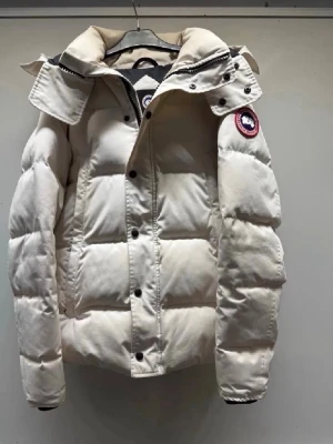 Vit dunjacka från Canada Goose - Snygg vit dunjacka från Canada Goose med klassisk patch på ärmen. Jackan har hög krage, stora knappar och dragkedja framtill. Puffig modell med breda kanaler och justerbar huva. Perfekt för kalla vinterdagar och riktigt varm.