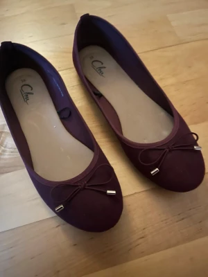 Vinröda ballerinaskor  - Snygga vinröda ballerinaskor med rund tå och dekorativ rosett framtill. Skorna har en klassisk och stilren design med små metallhylsor på rosetterna. 