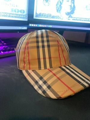 Burberry keps - Snygg keps från Burberry i beige med det ikoniska rutmönstret i svart, vitt och rött. Kepsen har böjd skärm och justerbar passform. Tillverkad i bomull och polyester för en bekväm känsla. Perfekt accessoar för att lyfta din streetwear-look samt perfekt ifall du vill sticka ut i sommar.