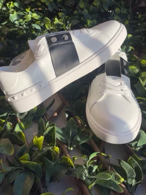 Valentino vita sneakers med svart detalj - Snygga vita sneakers från Valentino med bred svart läderdetalj över snörningen och silverfärgade nitar på sidan av sulan. Klassisk låg modell med vita skosnören och rund tå. Perfekta för dig som gillar stilrena och exklusiva skor.
