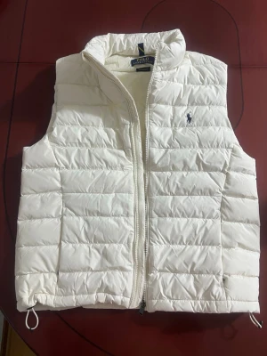 Vit dunväst från Polo Ralph Lauren - Stilren vit dunväst från Polo Ralph Lauren med klassisk quiltad design och dragkedja framtill. Västen har hög krage och ikoniska loggan broderad på bröstet. Perfekt för lager-på-lager och håller dig varm under kyliga dagar.