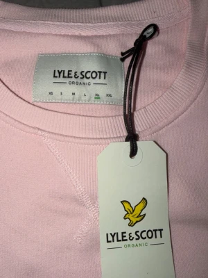 Ljusrosa sweatshirt från Lyle & Scott - Ljusrosa sweatshirt från Lyle & Scott i ekologisk bomull. Klassisk rund halsringning, ribbade muddar och ikonisk gul logga på bröstet. Enkel och stilren design som passar perfekt till jeans eller mjukisbyxor.