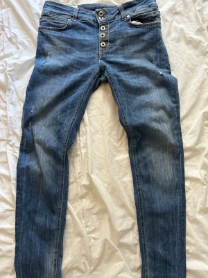 Dondup jeans - Snygga blå jeans med flera silverfärgade knappar framtill och lätt slitna detaljer på låren. Jeansen har klassisk femficksdesign och smal passform. Perfekta för en avslappnad och trendig look. Storlek: 30