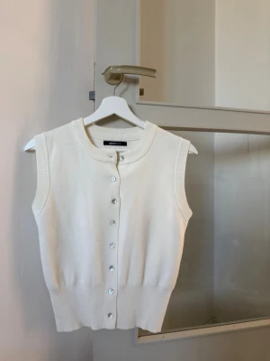 Vit stickad väst från Gina Tricot - Snygg vit stickad väst från Gina Tricot med rund halsringning och knappar framtill. Västen är ärmlös och har en ribbad kant nedtill för en clean look. Perfekt att styla över en skjorta eller t-shirt för en trendig vibe.