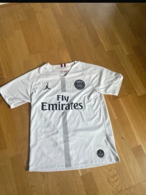 Vit PSG matchtröja Dembélé 10 - Snygg vit PSG matchtröja med Dembélé och nummer 10 på ryggen. Tröjan har Fly Emirates-tryck, PSG-logga och Jordan-logga framtill. Tillverkad i lätt och ventilerande material, perfekt för fotboll eller träning.