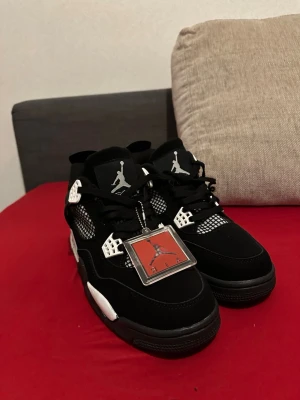 Svarta Air Jordan 4 Retro sneakers - Snygga Air Jordan 4 Retro sneakers i svart med vita detaljer och meshpaneler på sidorna. Skorna har klassisk Jumpman-logga på plösen och hängande tag. Ovandelen är i mocka och textil, sulan är vit och svart med synlig Air-enhet. Låda medföljer inte!